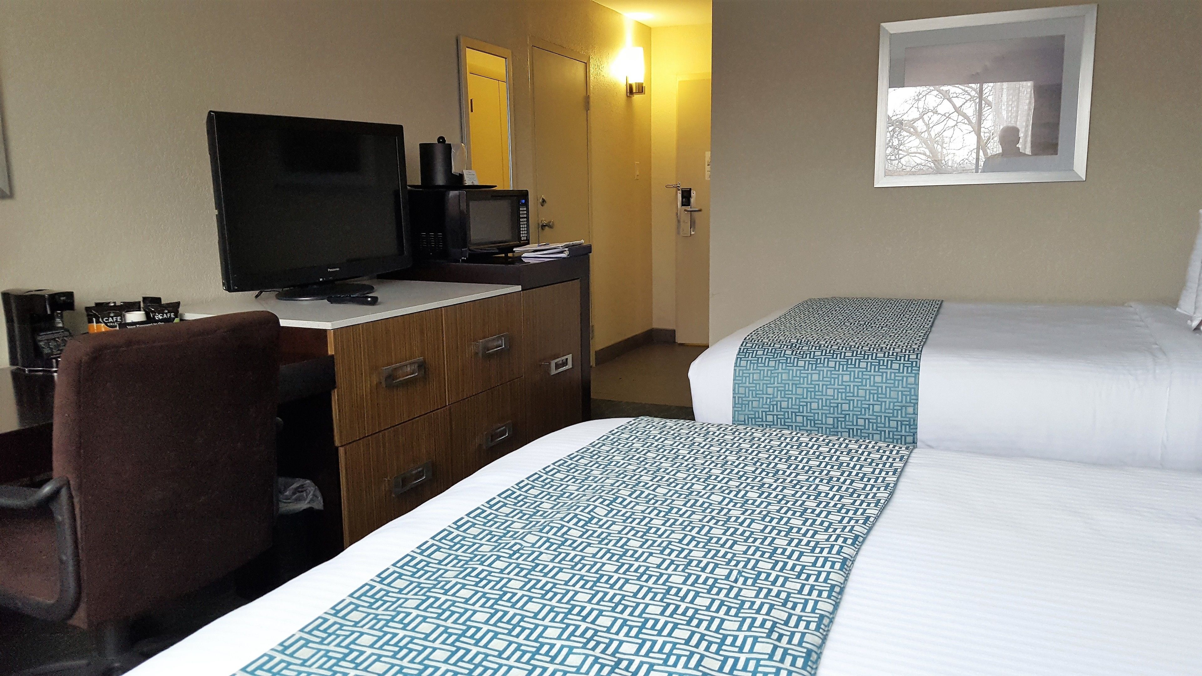 A hotel room with a desk, a television, a bed with patterned bedding, and a framed picture on the wall.