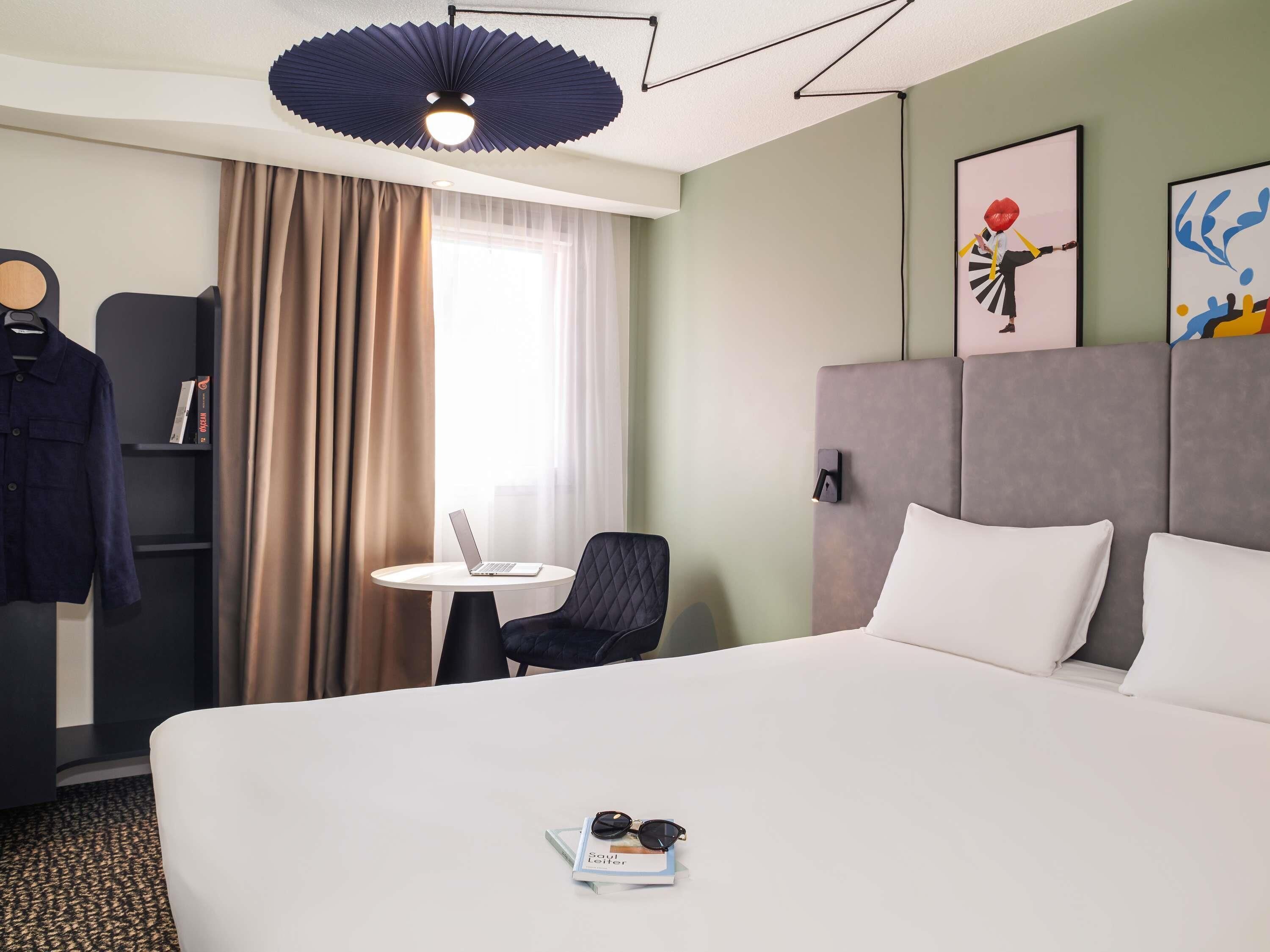 Ett hotellrum med en säng, ett skrivbord, en stol, en klädstång och ett fönster med gardiner.