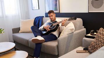 Iemand zit op een grijze bank en speelt gitaar, met een klein rond tafeltje en een smartphone op de vloer.