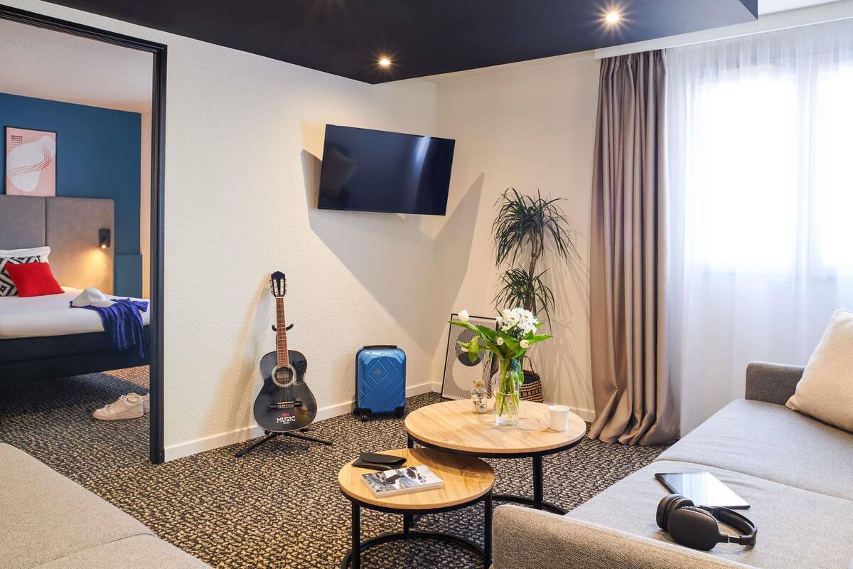 A hotel room with a bed, a TV, a guitar, a suitcase, a round table with a vase, and a sofa.