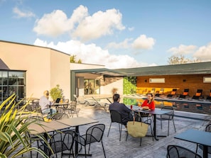 An outdoor patio with tables and chairs, a pool, and a modern building with large windows. - ibis Saint Nazaire Trignac (Trignac)