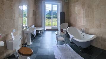 A bathroom with a toilet, sink, bathtub, and a small table with a chair. There is a view of a garden through a glass door.