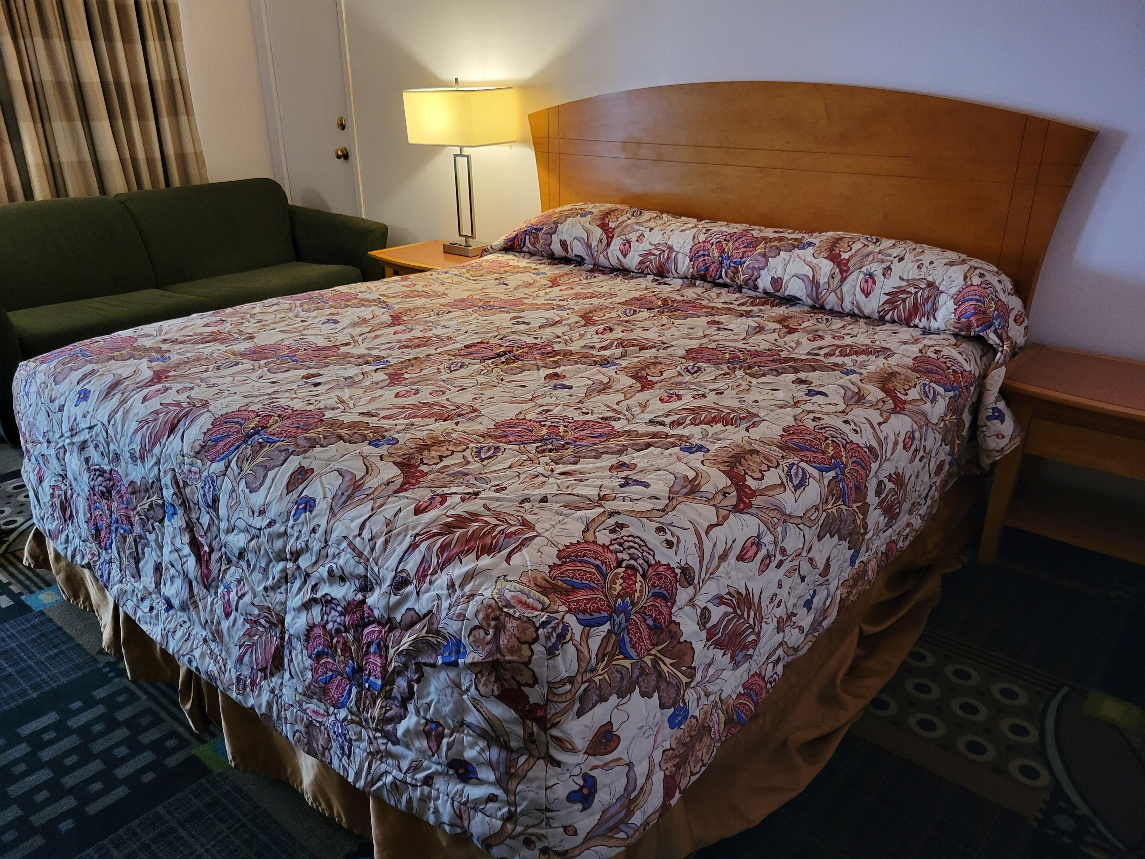 A hotel room with a large bed, a green sofa, a bedside table with a lamp, and a patterned bedspread.