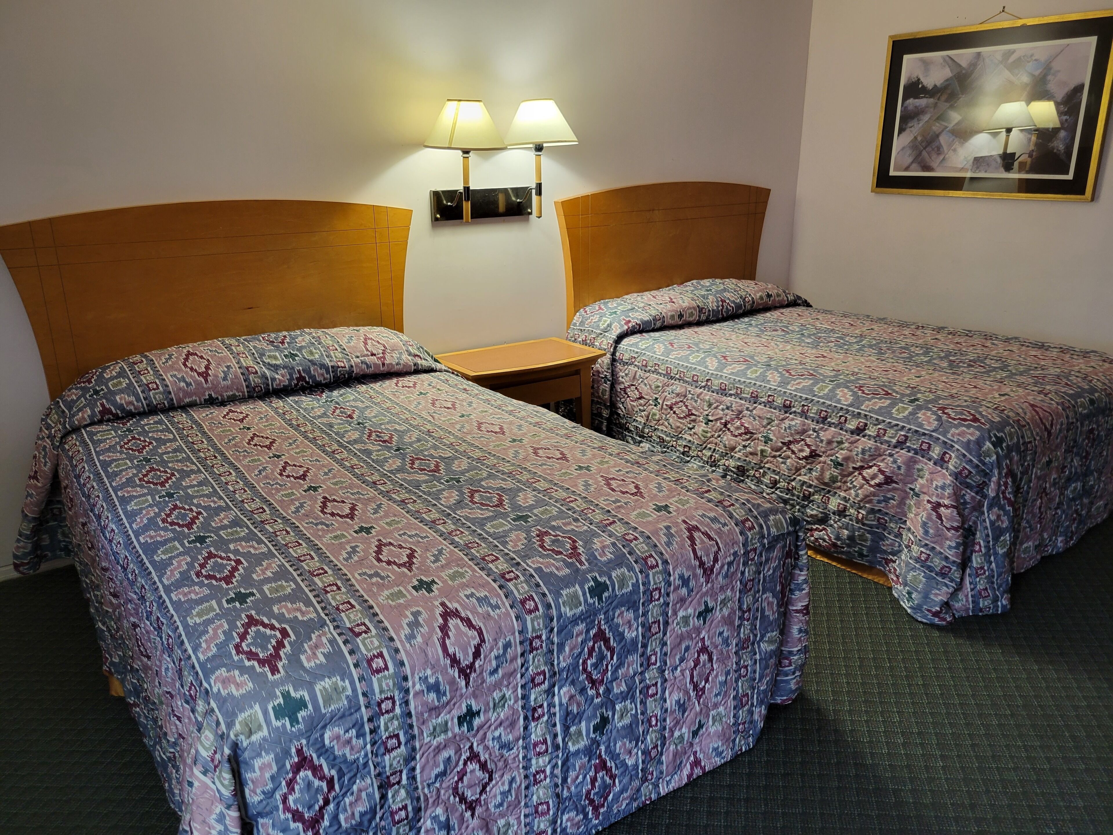 A hotel room with two beds, each with patterned bedding, a wooden headboard, a nightstand, and two wall-mounted lamps.