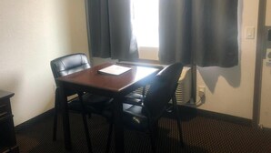 Quarto de hotel com mesa de madeira e quatro cadeiras pretas, janela com cortinas e aquecedor fixado na parede.