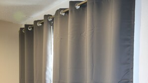 Quarto com janela coberta por cortinas cinzas, ar-condicionado branco fixado na parede e mesa de madeira com uma cadeira.