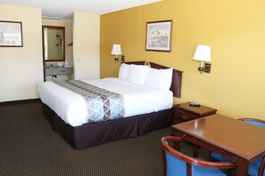A hotel room with a bed, bedside table, chair, desk, lamp, and a small table with a remote control.