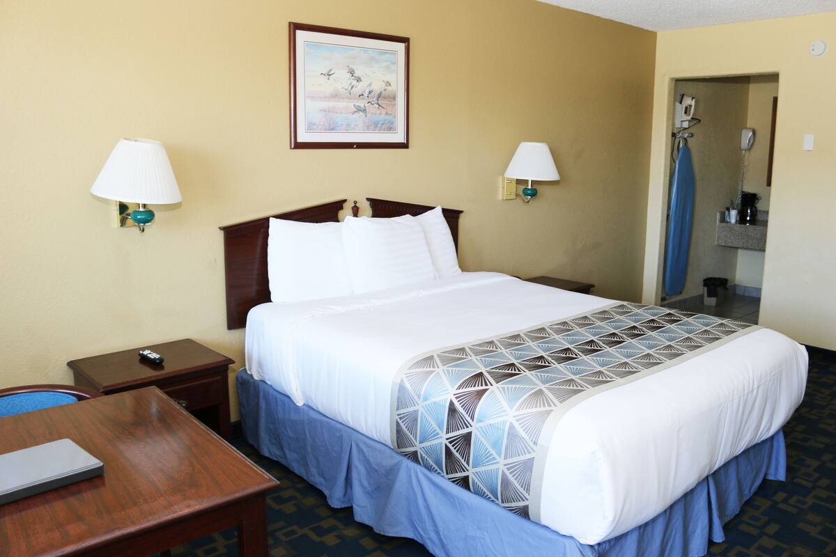 A hotel room with a bed, two bedside lamps, a painting on the wall, a desk with a laptop, and a bathroom visible through an open door.