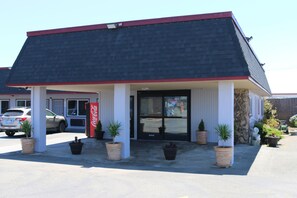A building with a dark roof and white walls, featuring a Coca-Cola sign, potted plants, and a parked car in front.