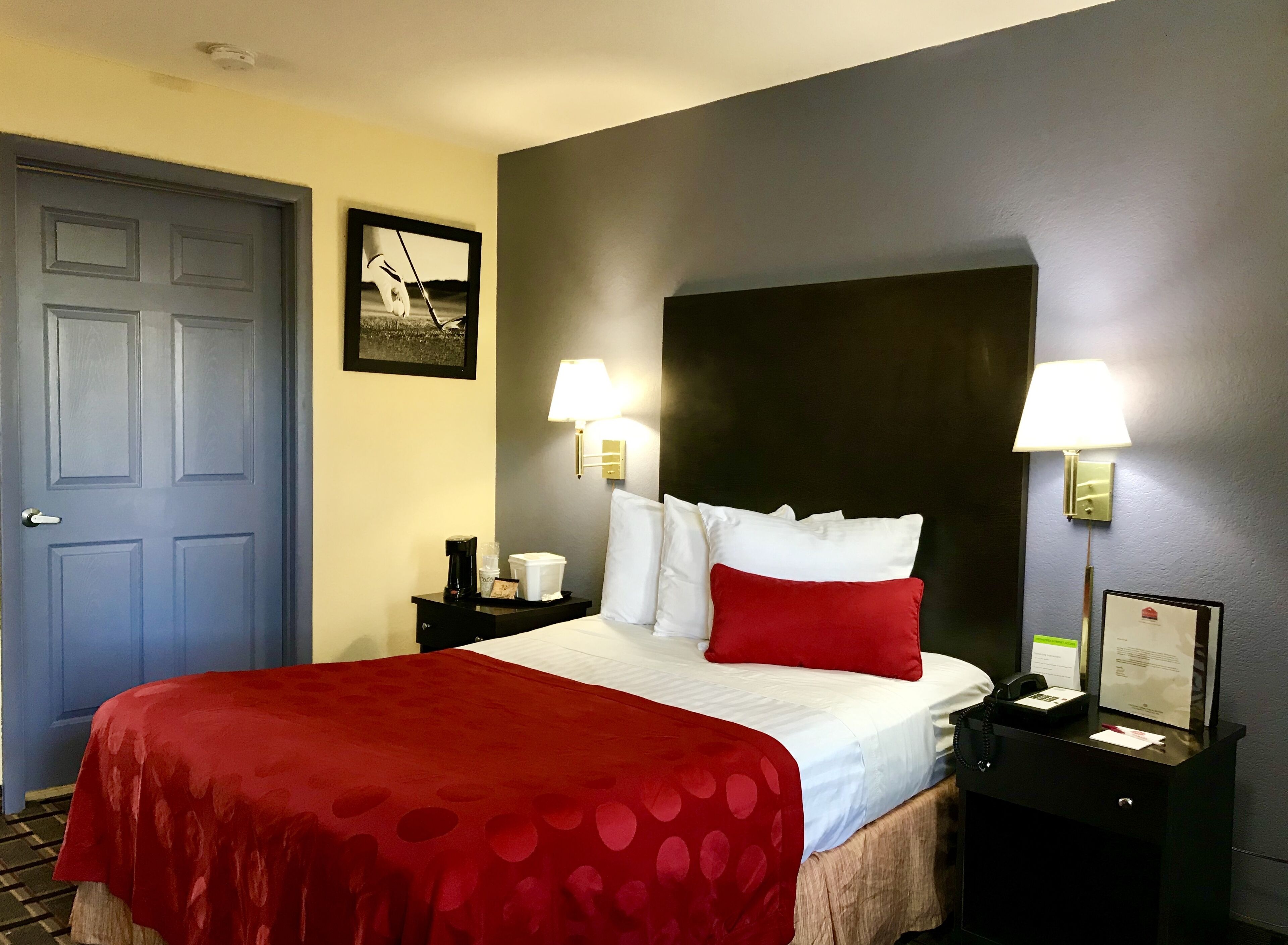 A hotel room with a bed, two bedside lamps, a nightstand with a phone and a lamp, a framed picture on the wall, and a door.