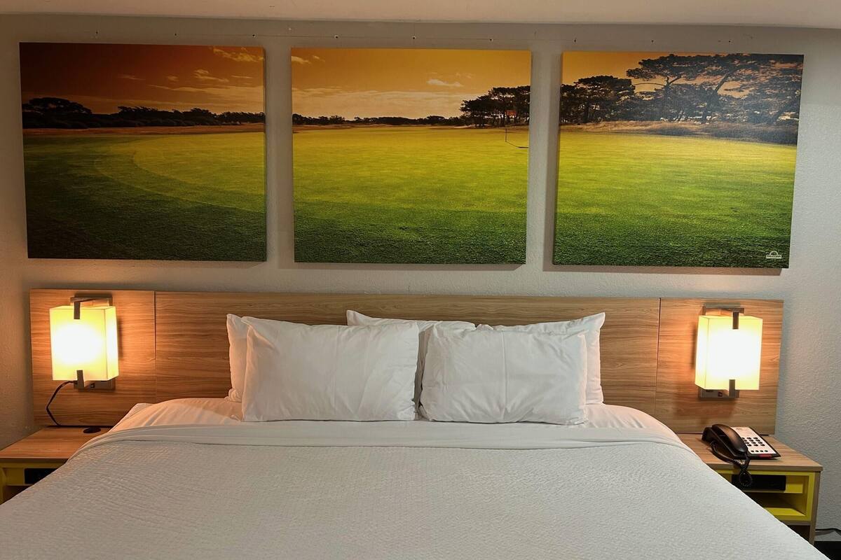 A hotel room with a bed, two bedside lamps, a nightstand with a phone, and three golf course paintings on the wall.