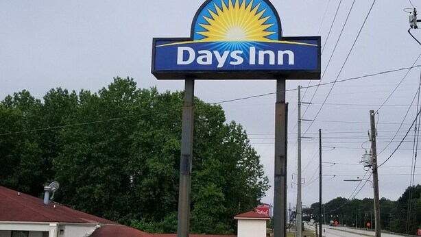 A Days Inn sign with a sun logo, a parking lot, and a building with a red roof. - Days Inn by Wyndham Augusta / Fort Eisenhower (Augusta)