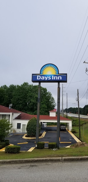 A Days Inn sign with a sun logo, a parking lot, and a building with a red roof. - Days Inn by Wyndham Augusta / Fort Eisenhower (Augusta)
