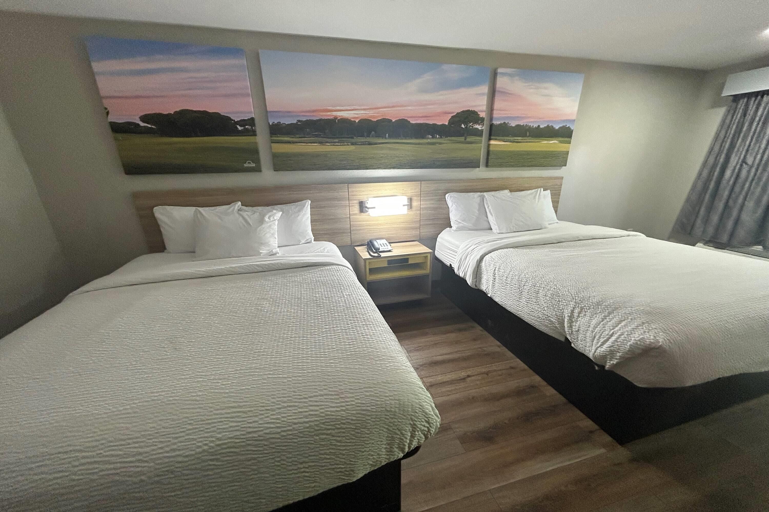 A hotel room with two beds, a nightstand with a lamp, and a large landscape painting above the beds.