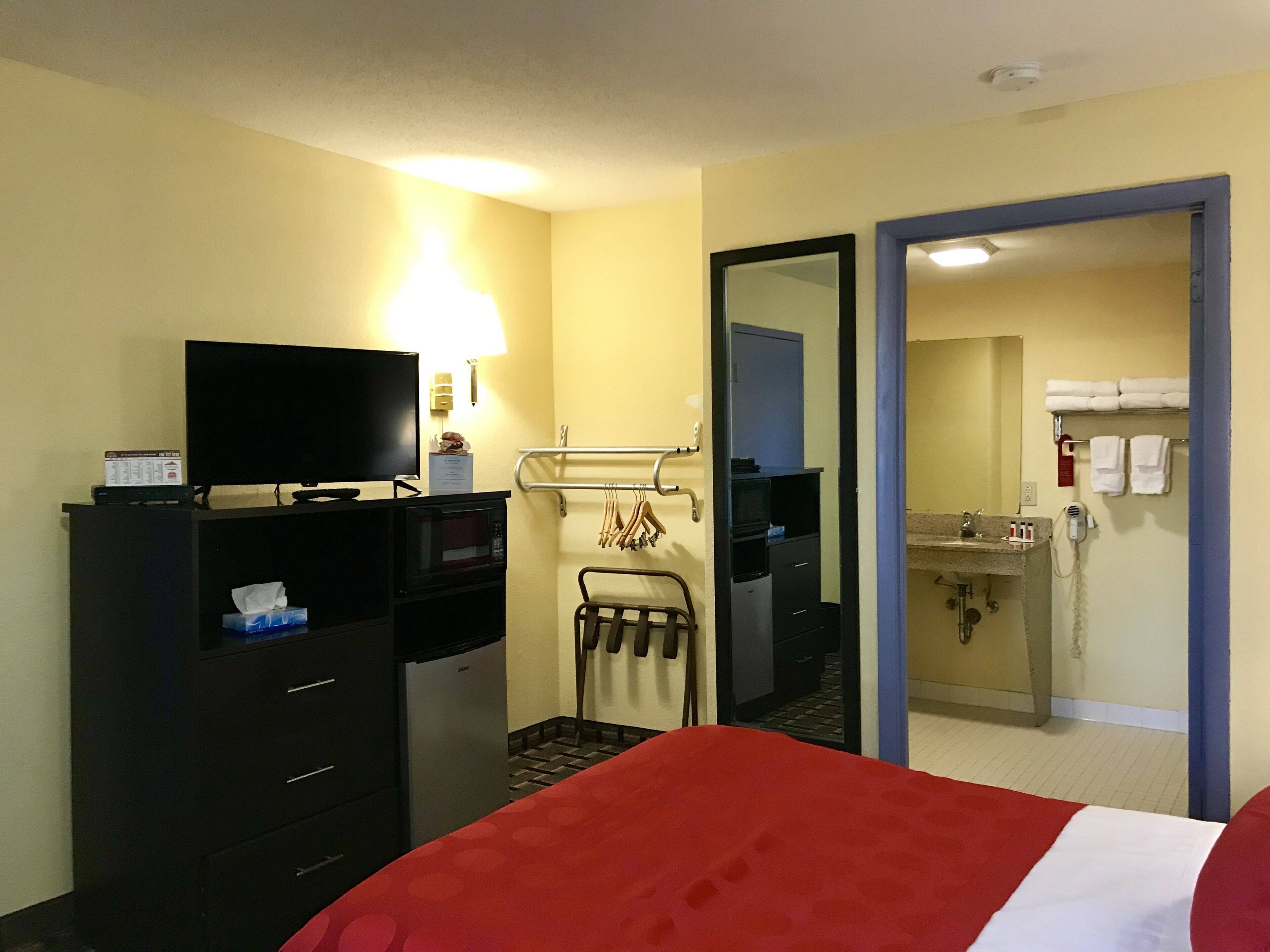 A hotel room with a bed, a TV, a desk with a computer, a microwave, a small trash bin, a mirror, a towel rack, and a bathroom with a sink and towel dispenser.
