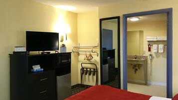 Habitación de hotel con cama, televisor, escritorio con computadora, microondas, basurero pequeño, espejo, toallero y baño con lavamanos y dispensador de toallas.