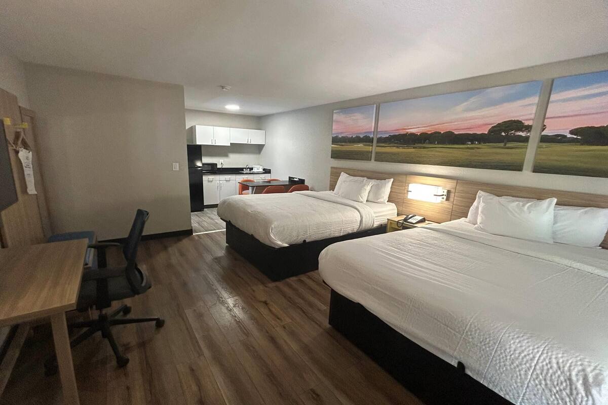 A hotel room with two beds, a desk, a chair, a small kitchenette, and a painting of a landscape.