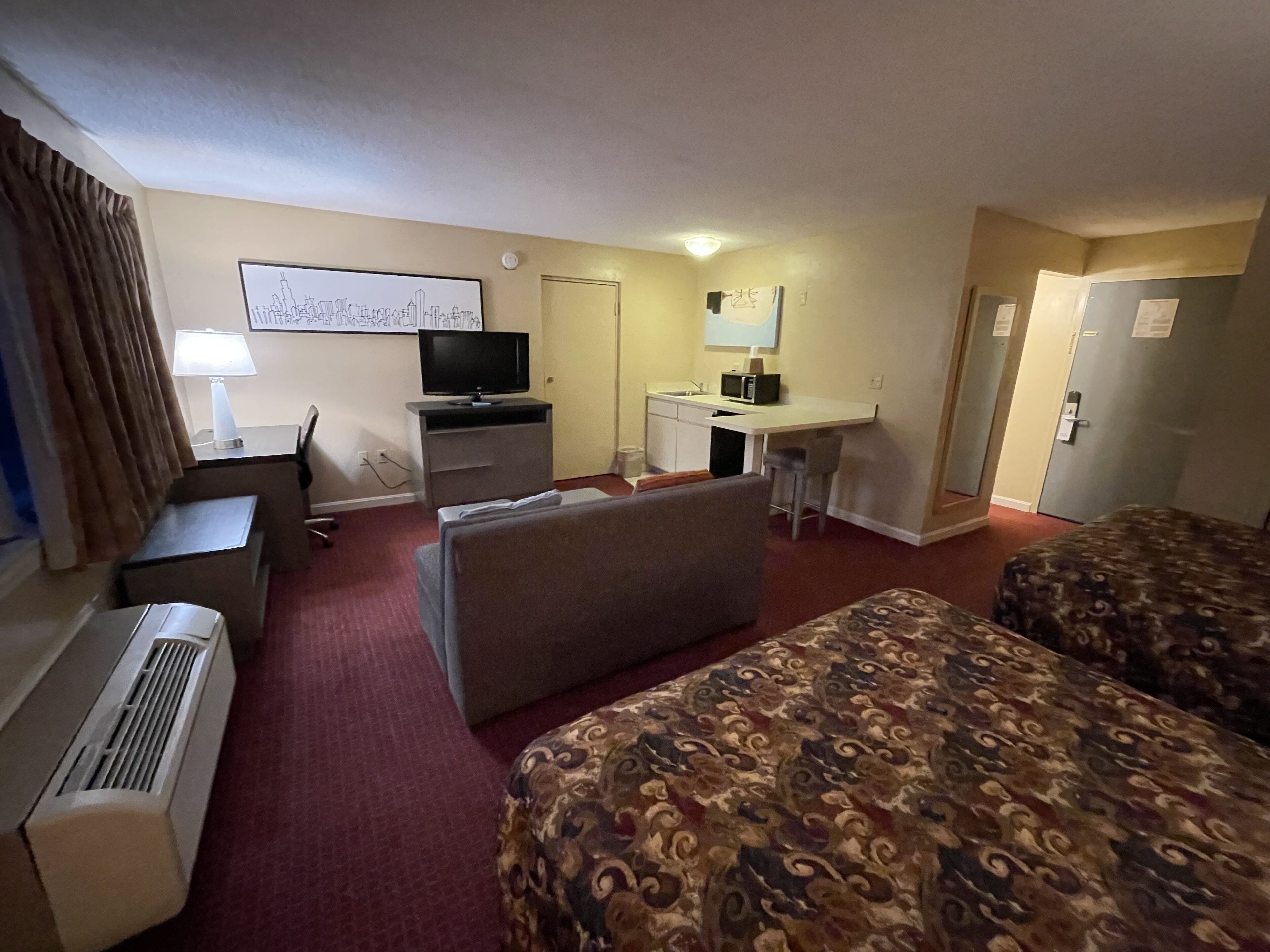 Quarto de hotel com cama, escrivaninha, televisão, pequeno espaço de cozinha e janela com cortinas.