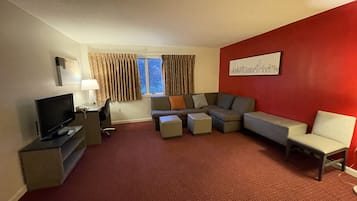 A hotel room with a red accent wall, a grey sectional sofa, a flat-screen TV, a desk with a lamp, and a window with curtains.