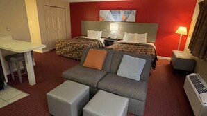 A hotel room with a red carpet, a grey sofa, two beds with patterned bedding, a desk, and a lamp.