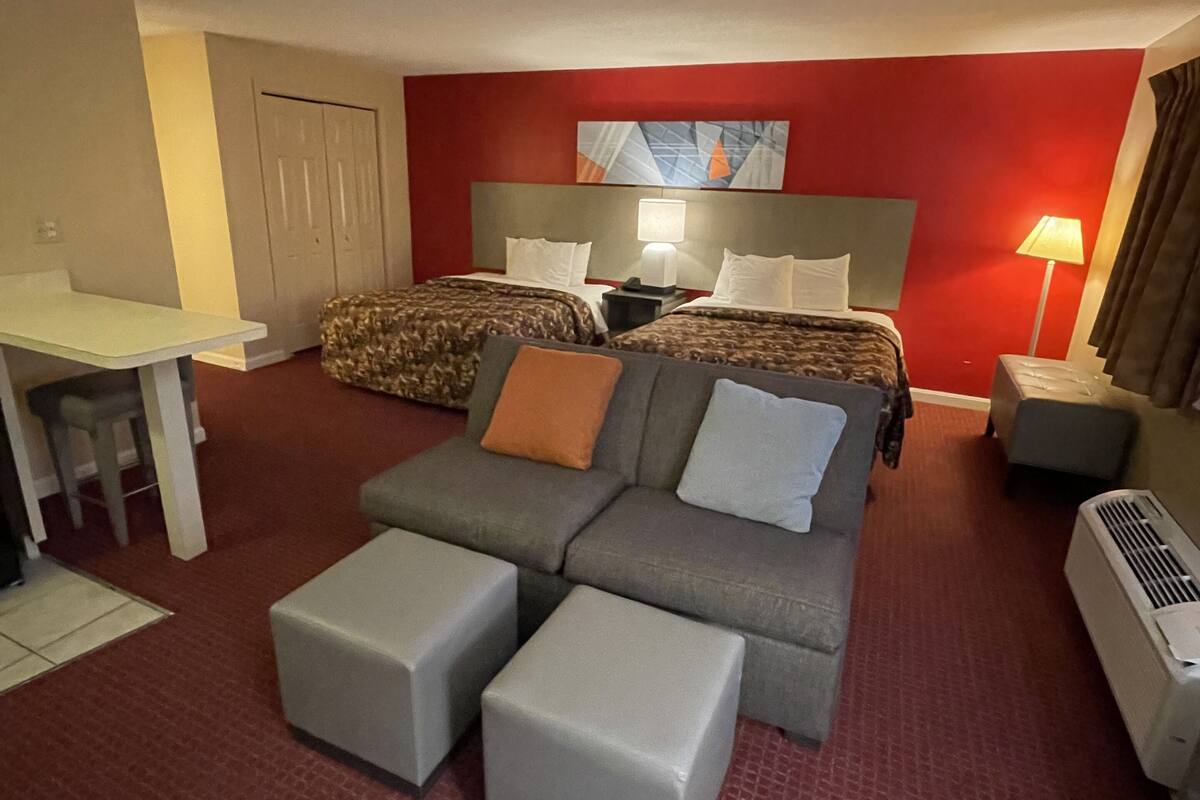 A hotel room with a red carpet, a grey sofa, two beds with patterned bedding, a desk, and a lamp.