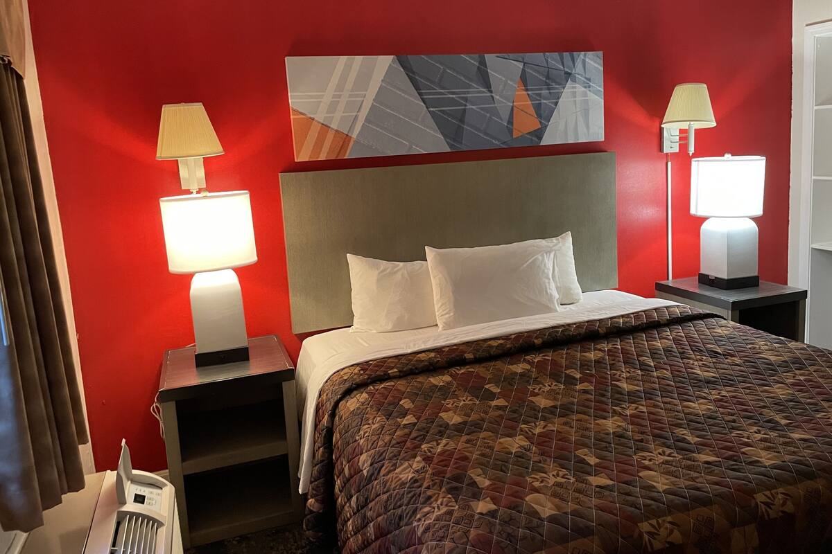 A hotel room with a red wall, a bed with white bedding, two bedside lamps, and a modern abstract painting above the bed.