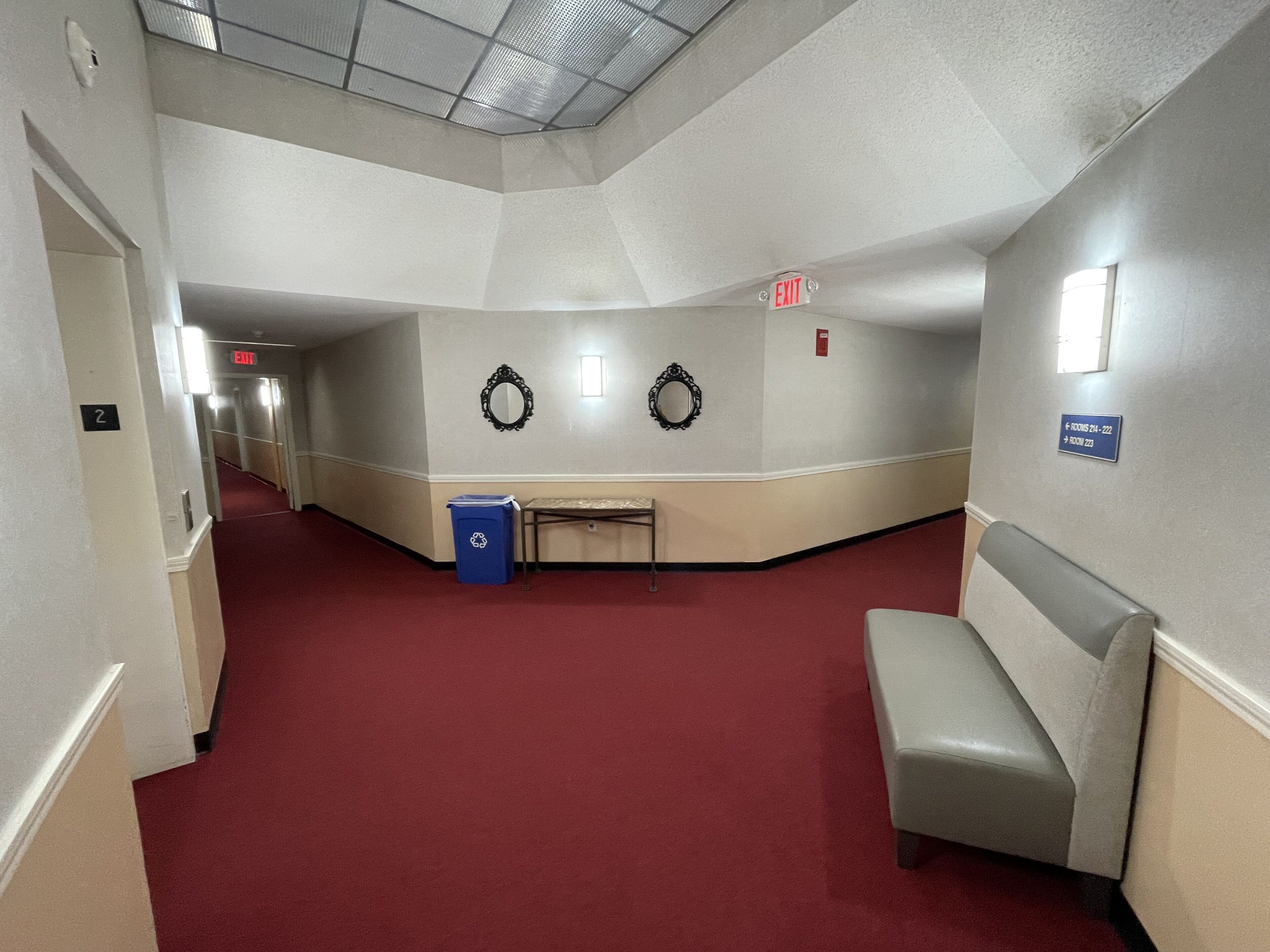 A hotel corridor with a red carpet, two wall-mounted mirrors, a blue trash bin, and a bench.