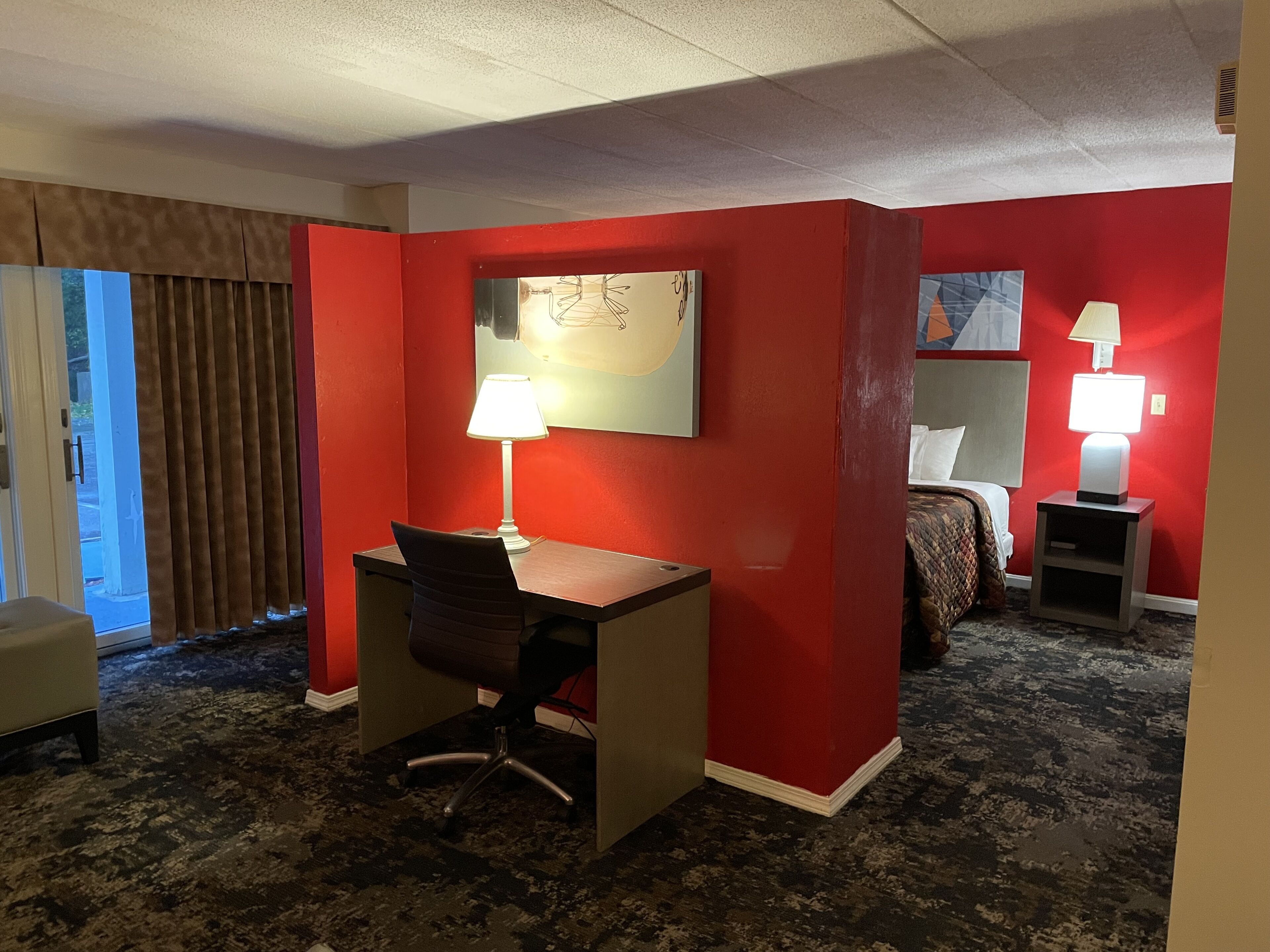 A hotel room with a desk, chair, two bedside lamps, a bed, and a large window with curtains.