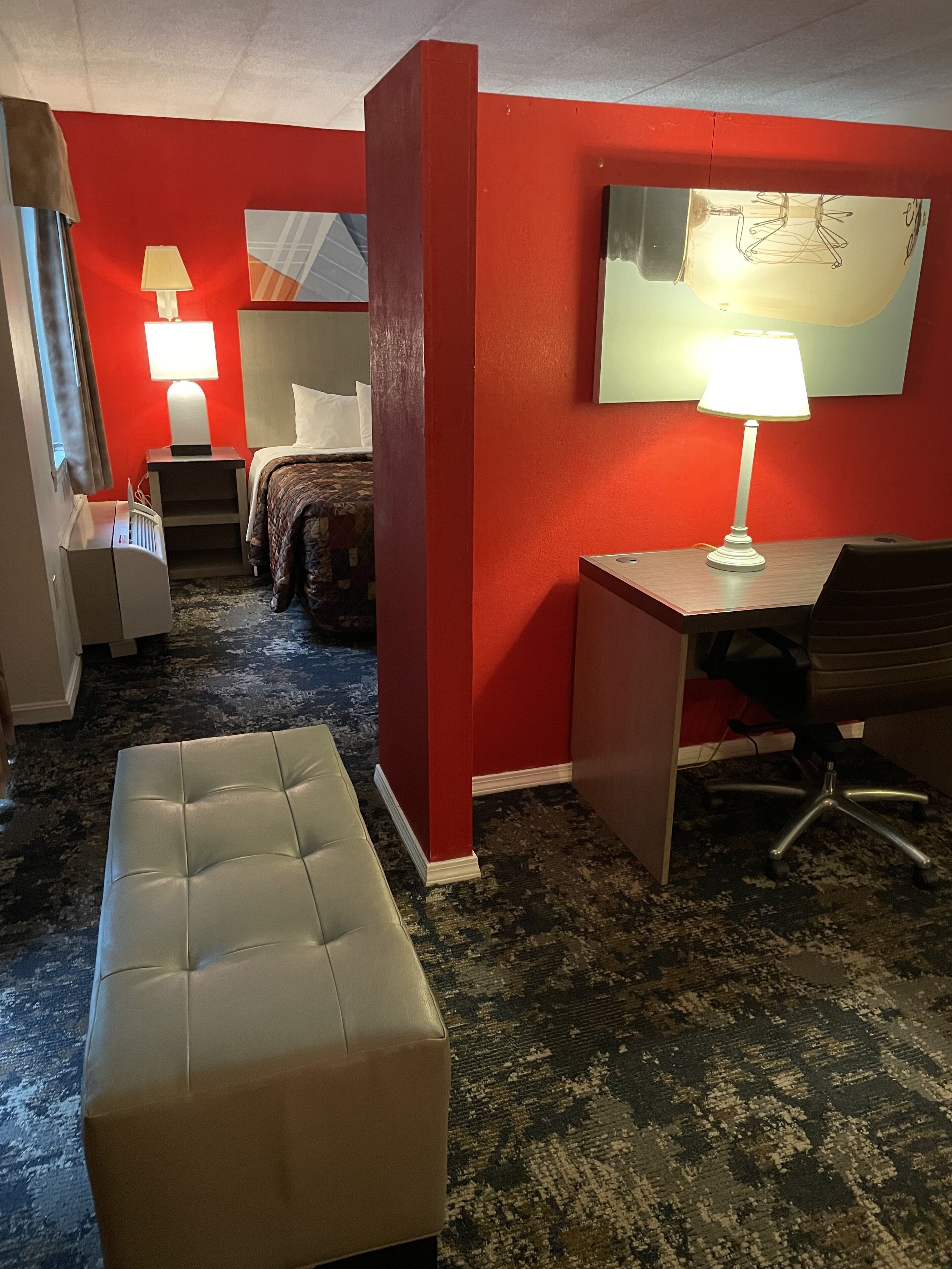 A hotel room with a red wall, a bed with a patterned bedspread, a desk with a lamp, a chair, and a cushioned bench.