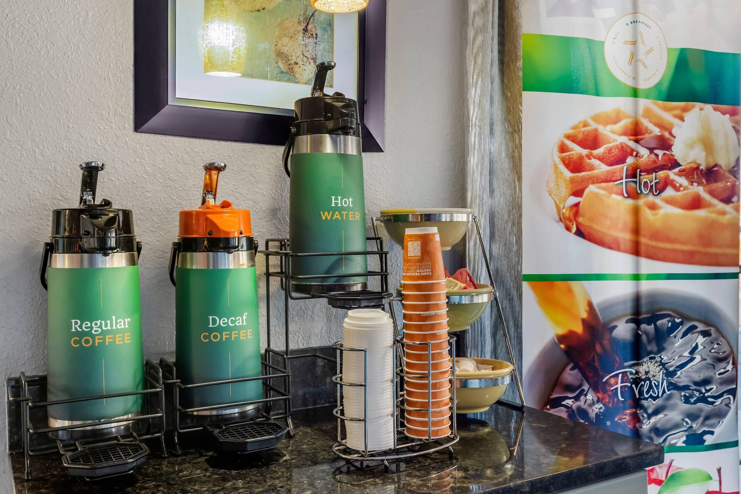 A coffee station with labeled dispensers for regular coffee and decaf coffee, a hot water dispenser, and a stack of disposable cups.