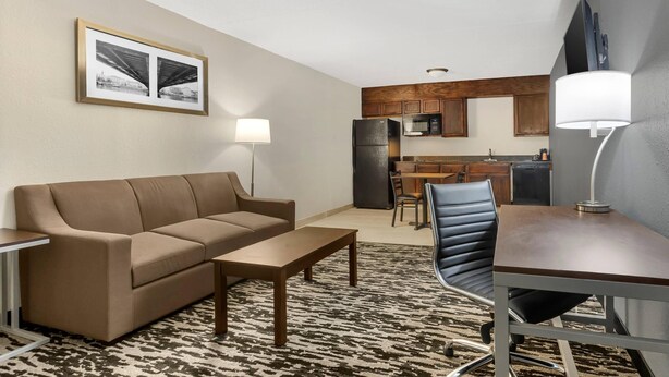 A modern living room with a sofa, coffee table, and a dining area with a table and chairs. A kitchen with a refrigerator and microwave is visible in the background. - Quality Inn & Suites Easley - Greenville (Easley)