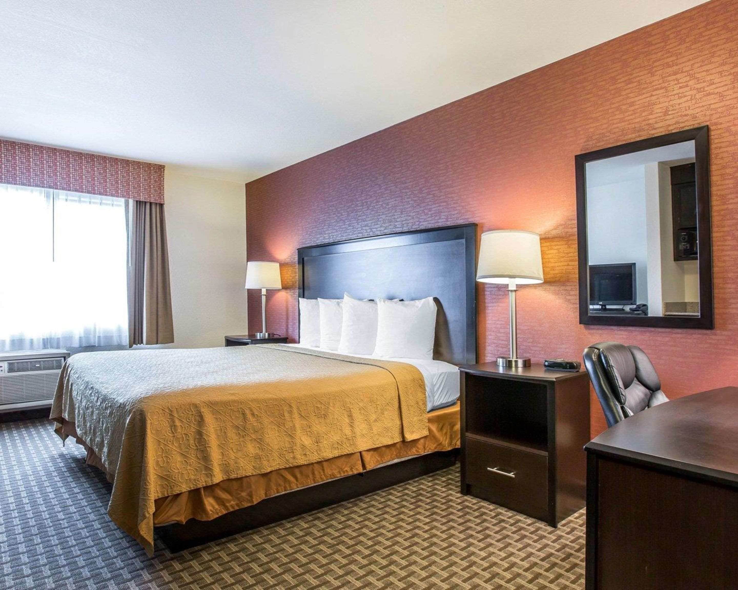 Quarto de hotel com uma cama grande, duas mesas de cabeceira com luminárias, uma escrivaninha com cadeira, uma televisão e um espelho na parede.