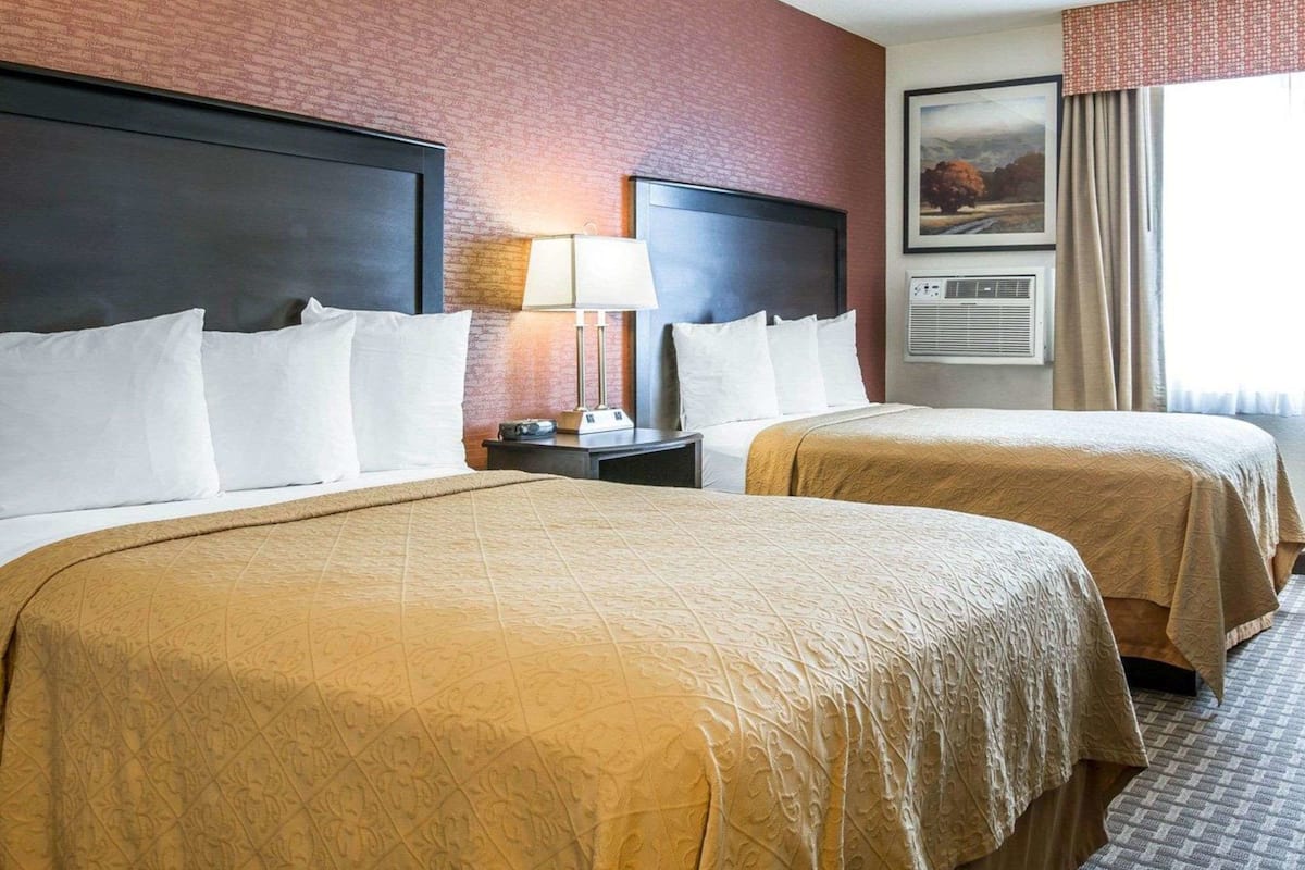 A hotel room with two beds, a nightstand with a lamp, a painting on the wall, and an air conditioning unit.