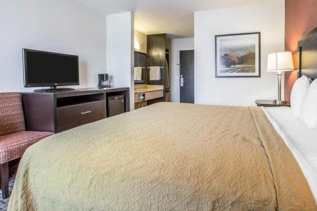 A hotel room with a bed, a desk with a television, a chair, a desk lamp, and a painting on the wall.