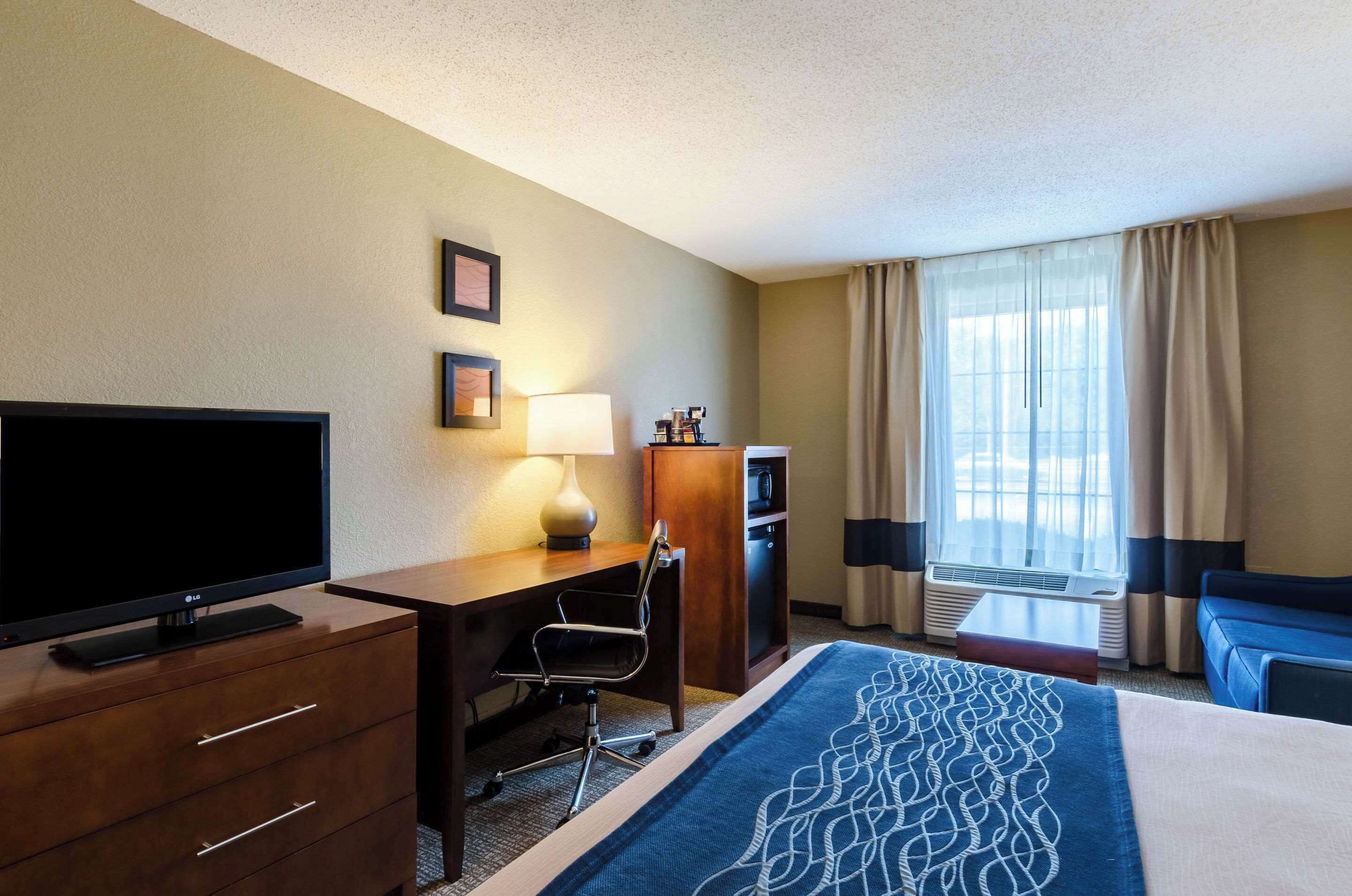 A hotel room with a bed, a desk with a chair, a television, a lamp, a minibar, and a window with curtains.