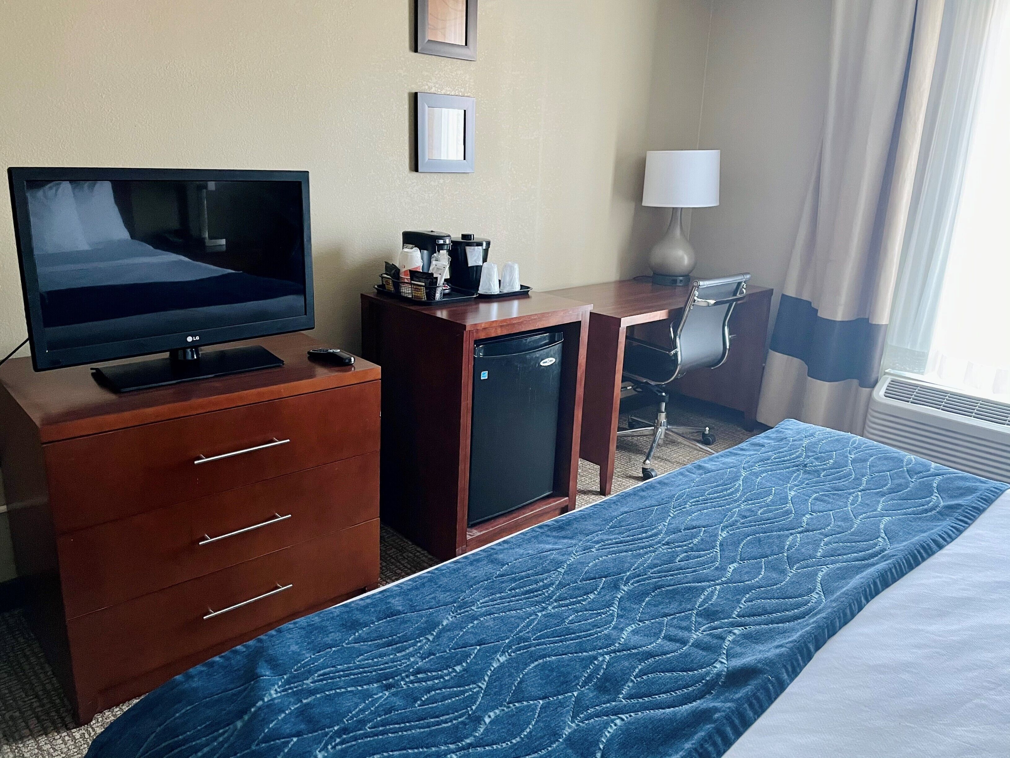 A hotel room with a bed, a desk with a computer, a television, and a refrigerator.