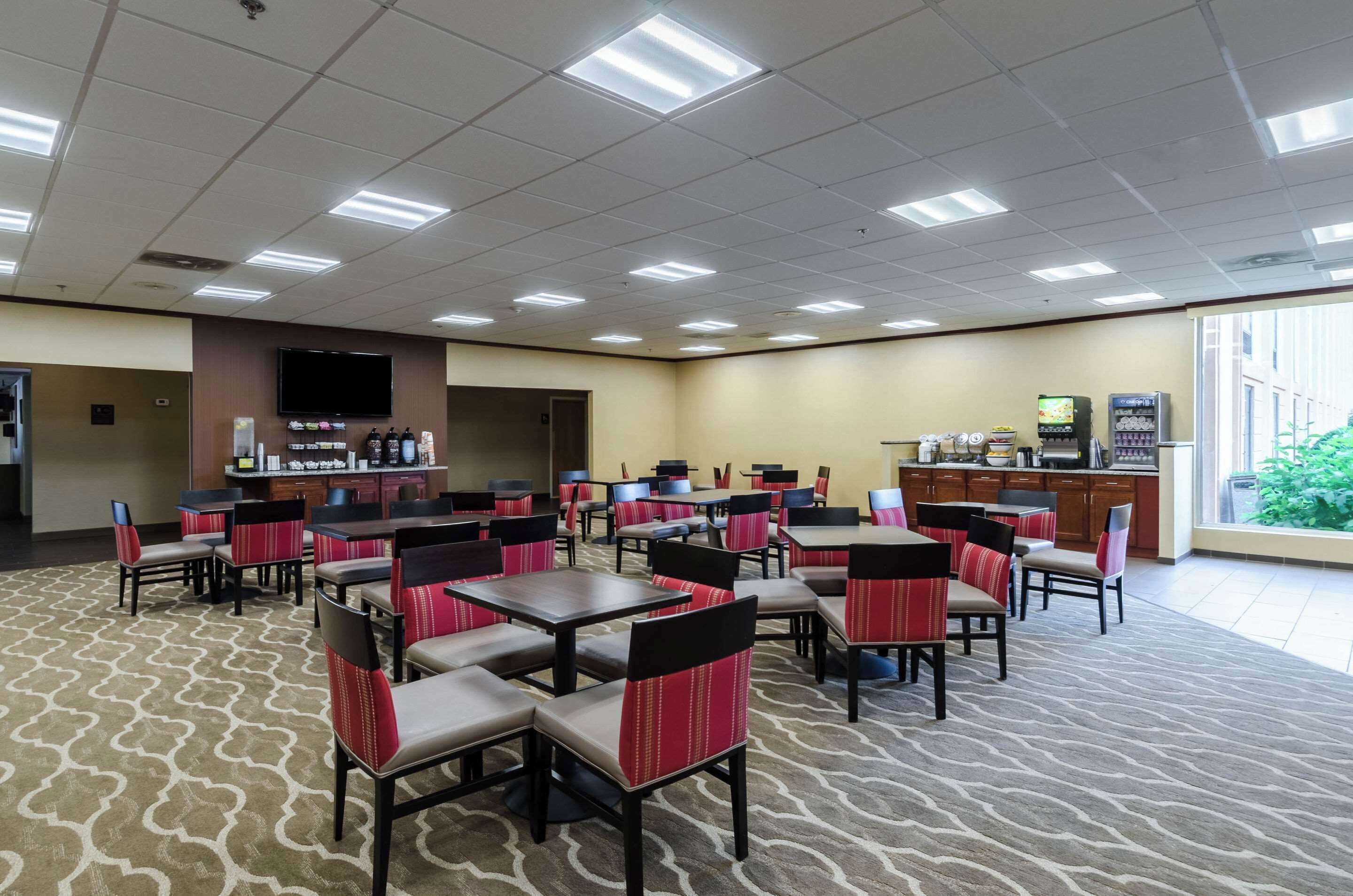 A hotel dining area with multiple tables and chairs, a kitchen area, and a large window providing a view of the outdoors.