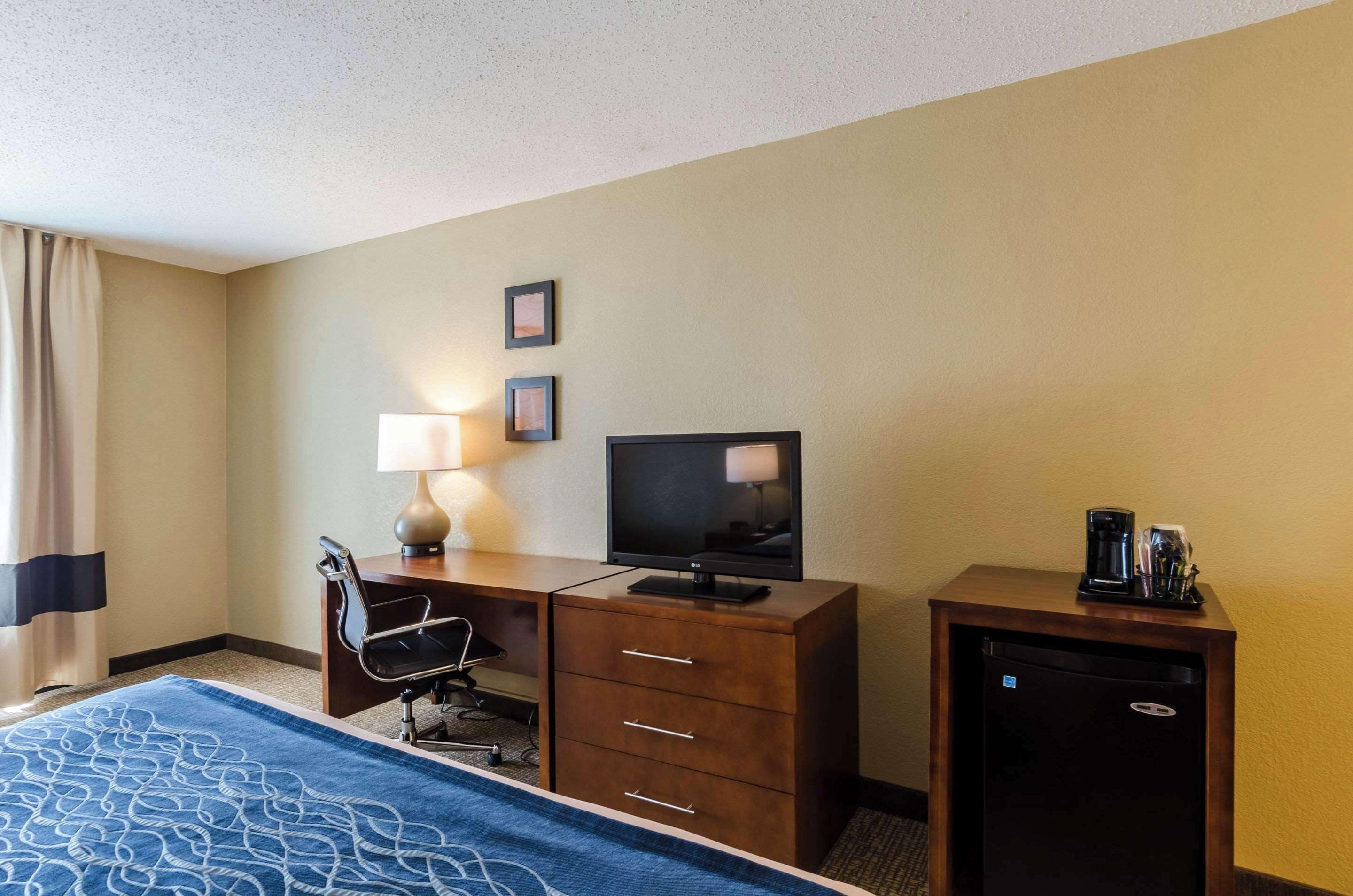 Habitación de hotel con cama, escritorio con lámpara, televisor, cafetera y refrigerador pequeño.