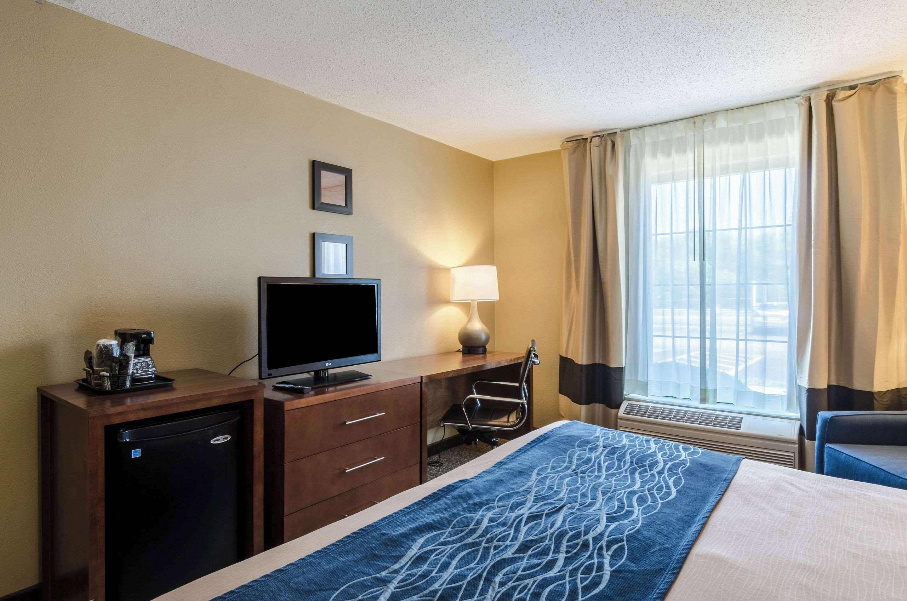Una habitación de hotel con cama, un escritorio con computadora, cafetera, lámpara y ventana con cortinas.