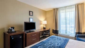 Una habitación de hotel con cama, un escritorio con computadora, cafetera, lámpara y ventana con cortinas.