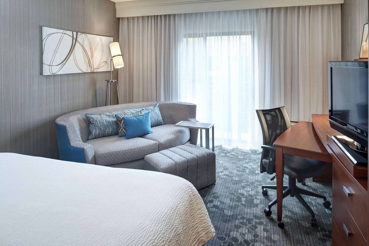 A hotel room with a large bed, a sofa, a desk with a TV, a lamp, and a view of the city through the window.