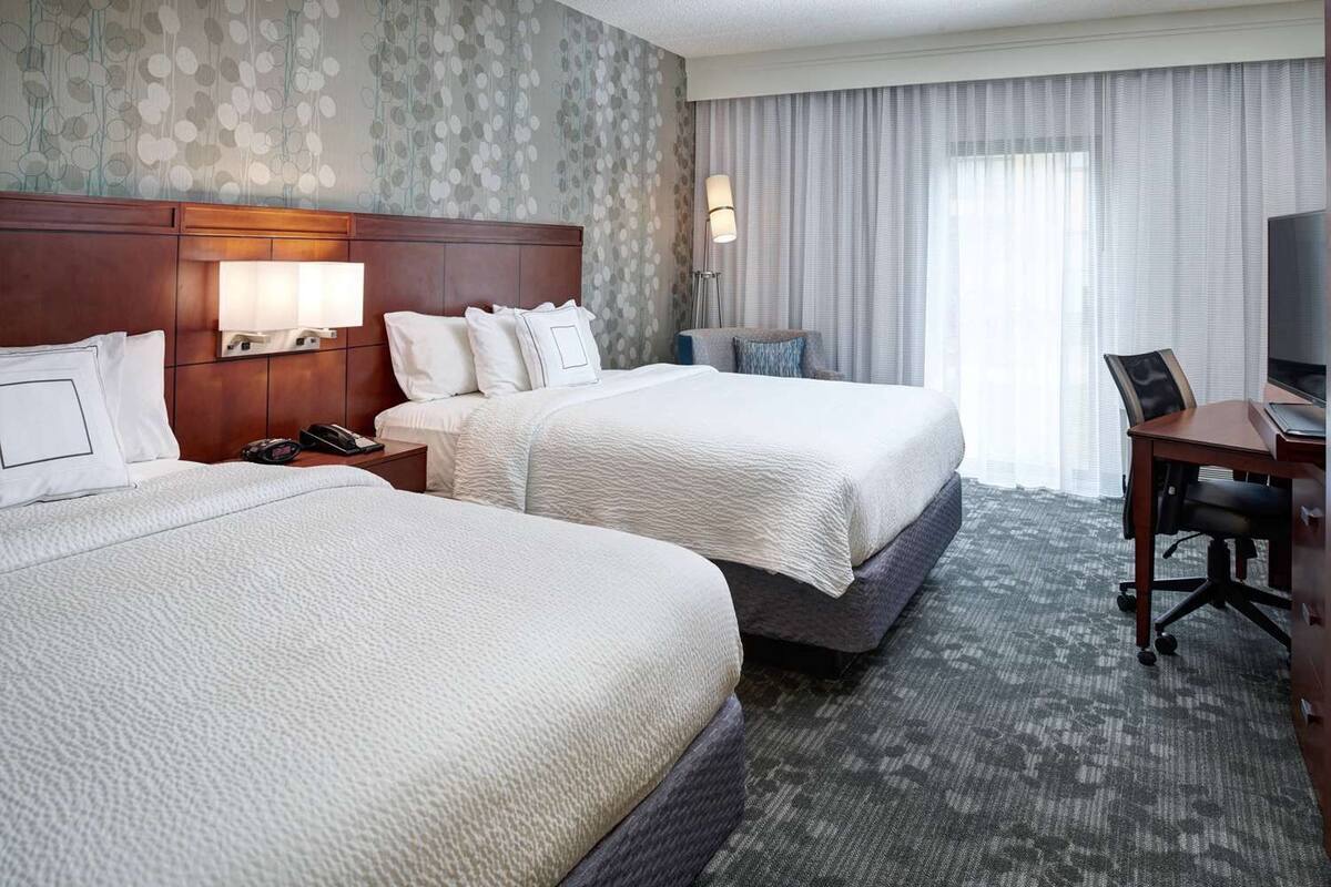 A hotel room with two beds, a desk, a chair, a lamp, and a television.