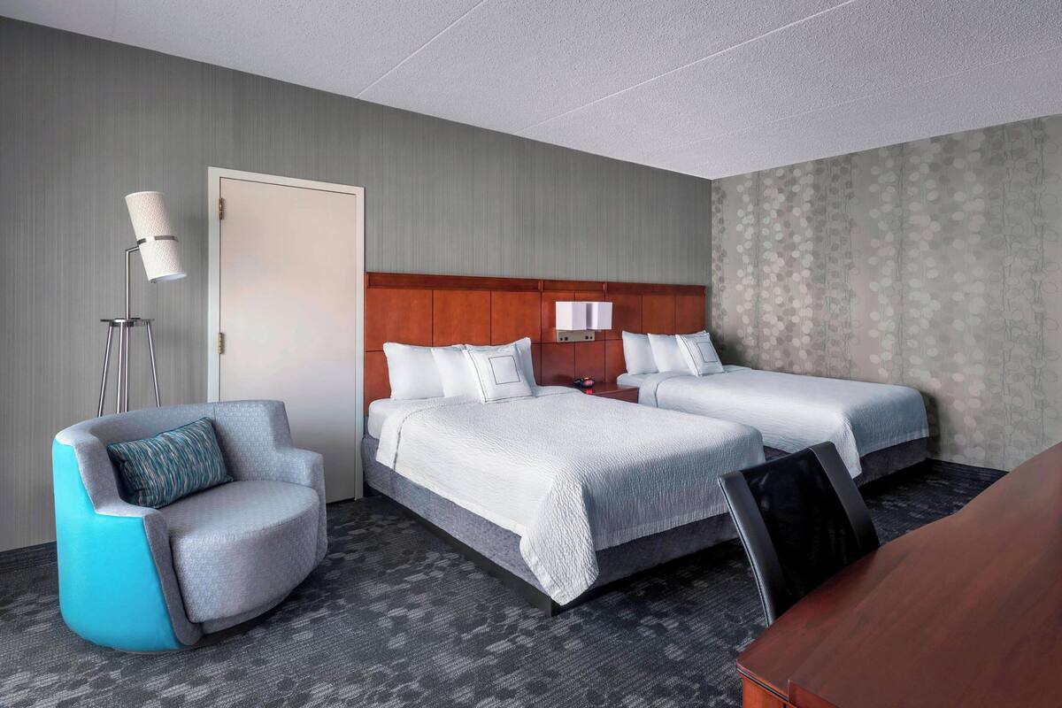 Habitación de hotel con dos camas, una zona de estar con una silla, un escritorio y una lámpara.