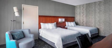 A hotel room with two beds, a seating area with a chair, a desk, and a lamp.
