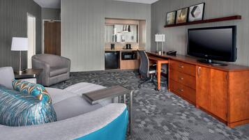 A modern hotel room with a grey sofa, a wooden desk, a flat-screen TV, a kitchenette, and a large mirror.