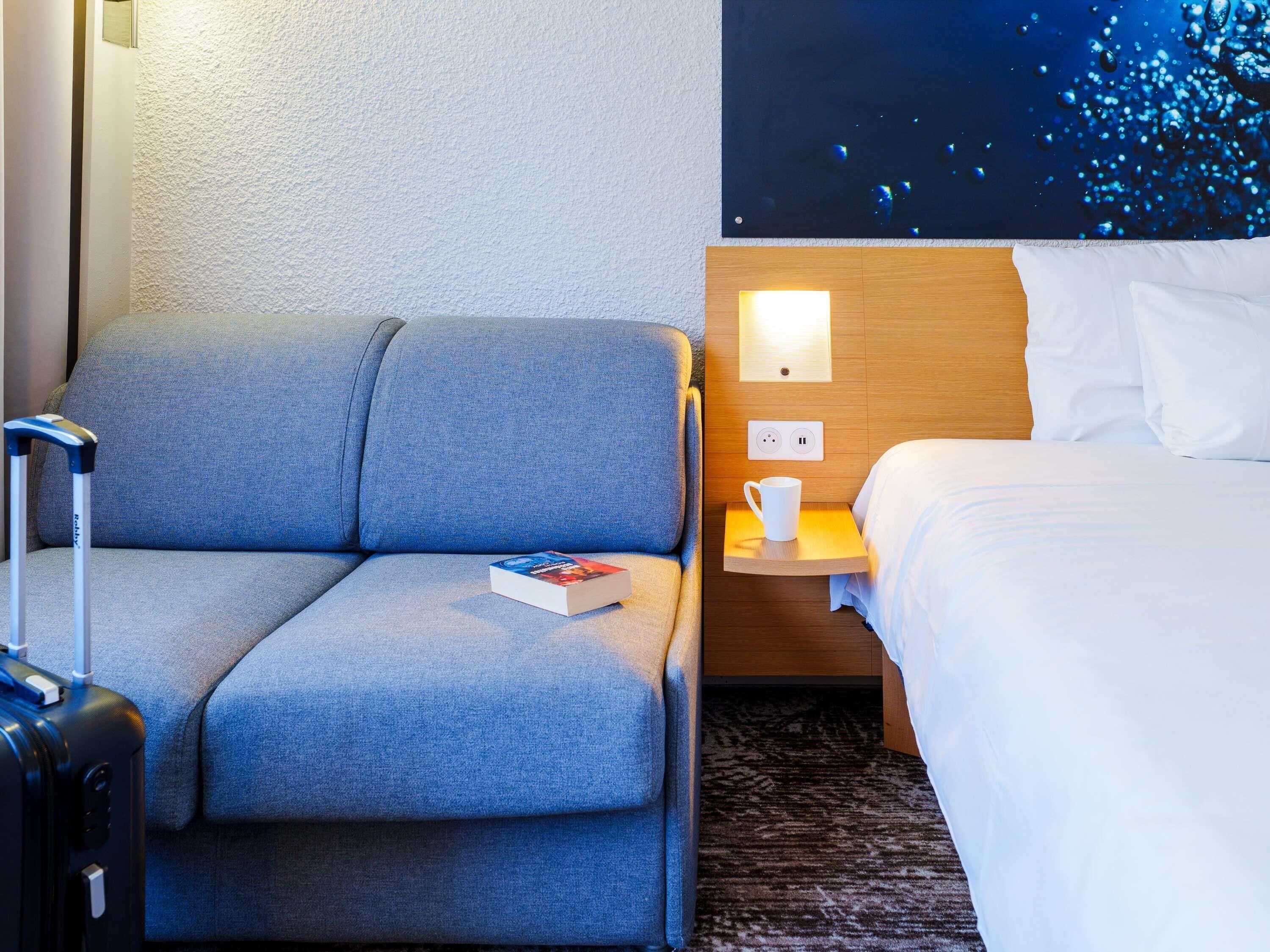 Habitación de hotel con un sofá gris, una mesita de noche pequeña con una taza blanca, una cama con ropa de cama blanca y un televisor de pared que muestra una imagen abstracta azul.