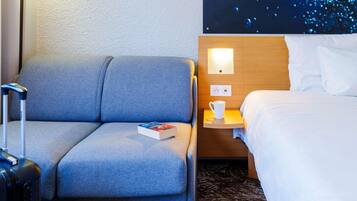 Habitación de hotel con un sofá gris, una mesita de noche pequeña con una taza blanca, una cama con ropa de cama blanca y un televisor de pared que muestra una imagen abstracta azul.