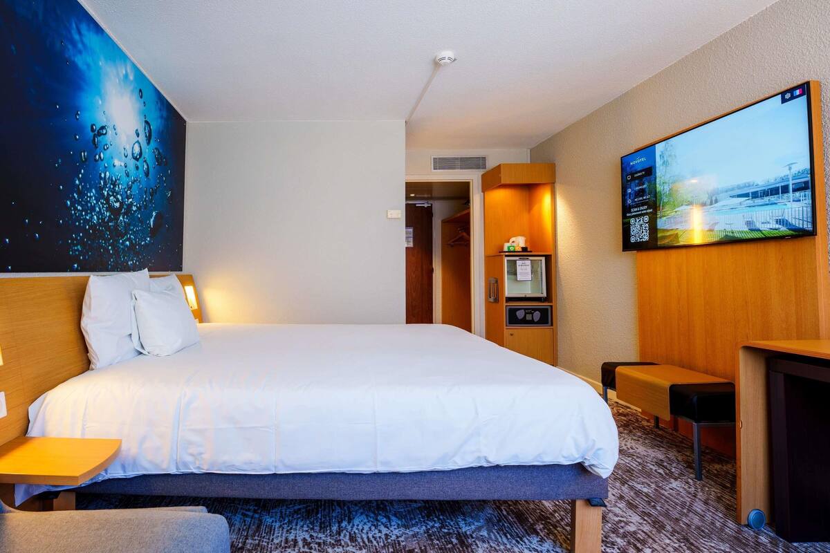A hotel room with a large bed, a TV, a desk, and a painting on the wall.