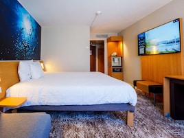 A hotel room with a large bed, a TV, a desk, and a painting on the wall.