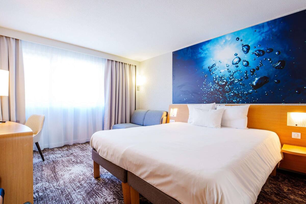 A hotel room with a large bed, a desk, a chair, a wall mural, and a window with curtains.